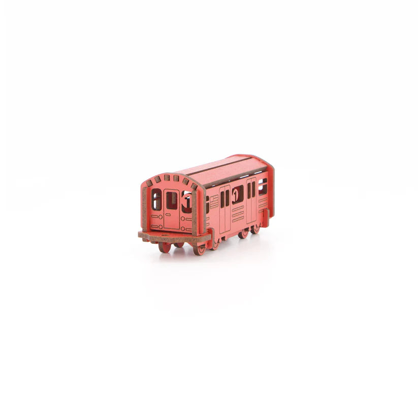 A small, salmon-pink wooden model shaped like a classic New York City subway car. The body is long and rectangular with smooth edges and simple cut‑out details suggesting windows and doors. The surface is flat and painted in a bold, uniform salmon-pink, giving it a clean, graphic feel. It stands upright on its base and captures the familiar silhouette of an NYC train in a compact, tactile form.