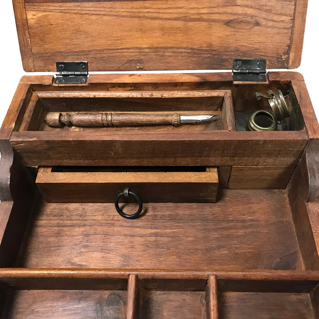The image shows a weathered, portable teak wood writing desk with its lid open, revealing interior storage compartments. Inside are a vintage‑style wooden pen and a small ink bottle topped with a distressed brass cap, reinforcing the desk’s antique, well‑traveled look.