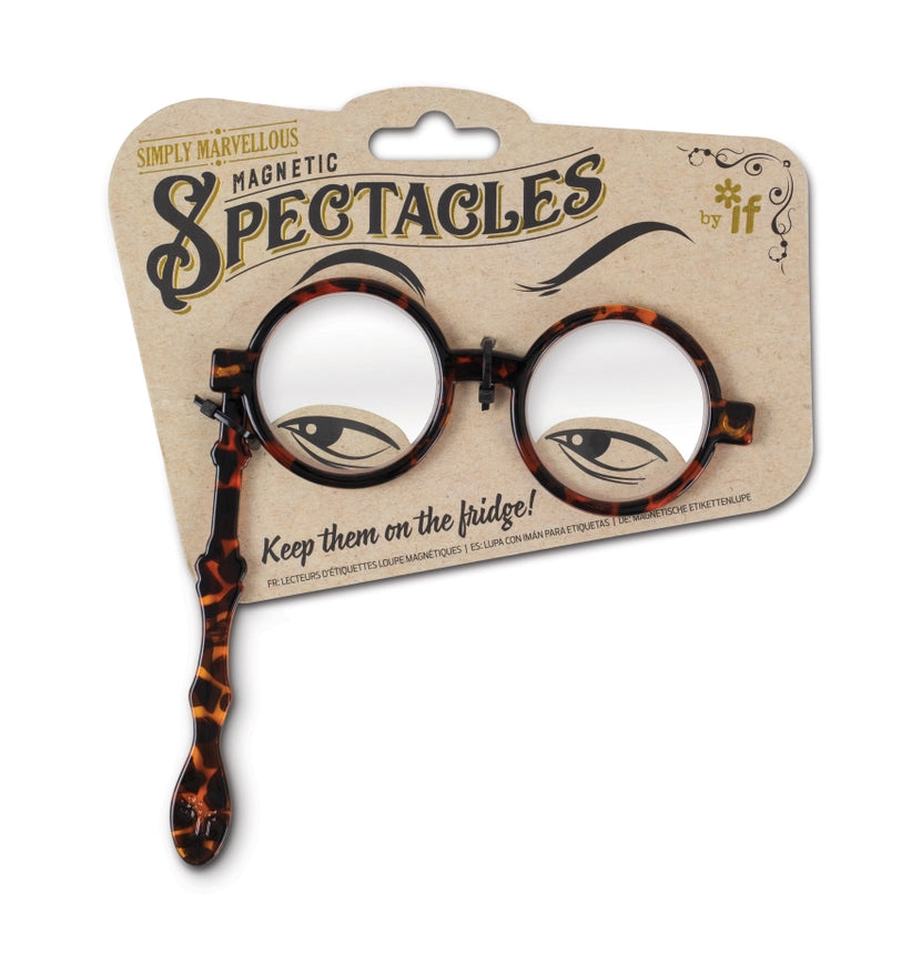 The image shows the Simply Marvellous Magnetic Spectacles with round, tortoiseshell‑patterned frames in a warm brown color palette. The lorgnette design includes a long side handle, making the glasses easy to recognize by touch. They are presented in compact “Try Me” packaging with an open front that allows handling without removing the glasses. The lenses contain hidden magnets, allowing the spectacles to stick to a fridge or other magnetic surface so they remain in a predictable, easy‑to‑find location.