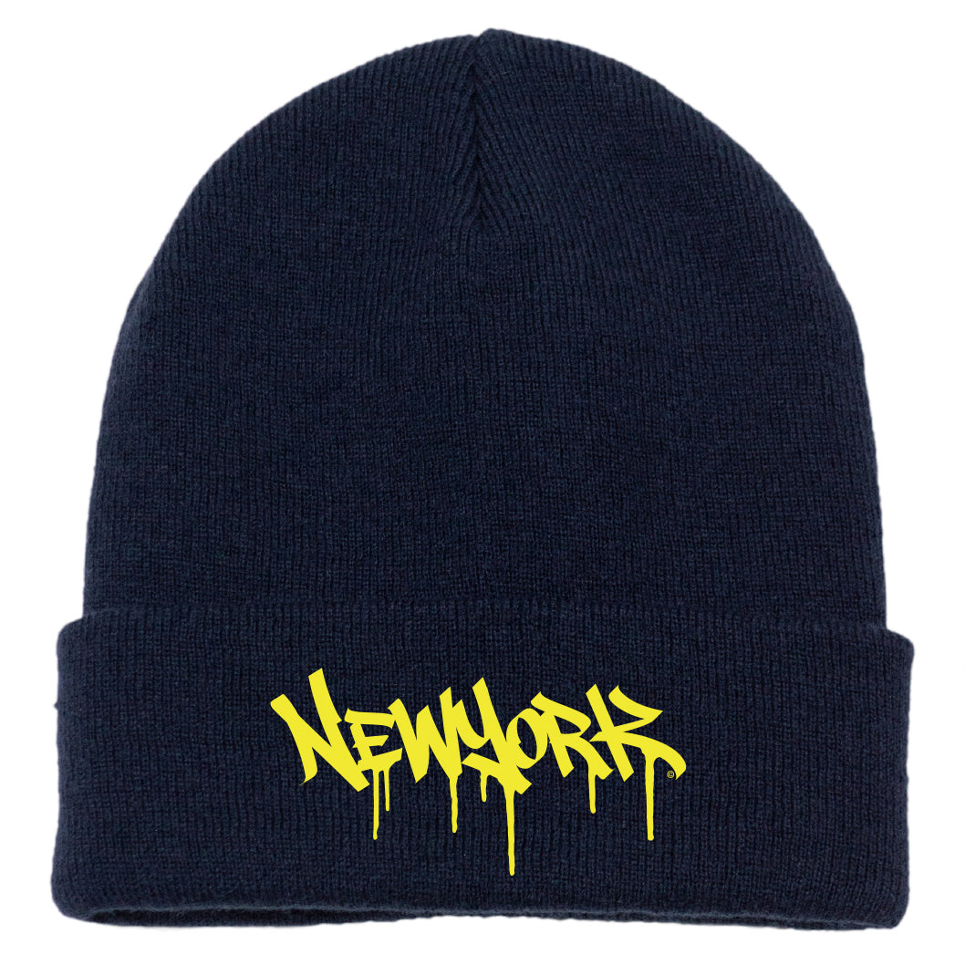 The image features a navy acrylic knit cap with a folded cuff, accented by bright yellow melting block-letter text spelling “New York.”