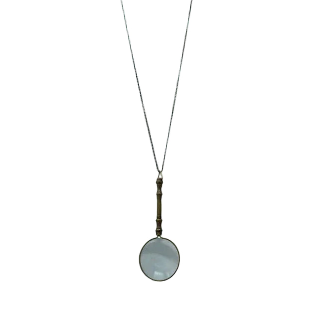 On a white background, the necklace displays an ornate magnifying glass pendant made of aged brass. The magnifying glass is about four inches long, with a decorative handle and a clear, working lens. It hangs from a matching aged brass scroll chain and closes with a small lobster clasp, giving the piece a refined, antique-inspired appearance.