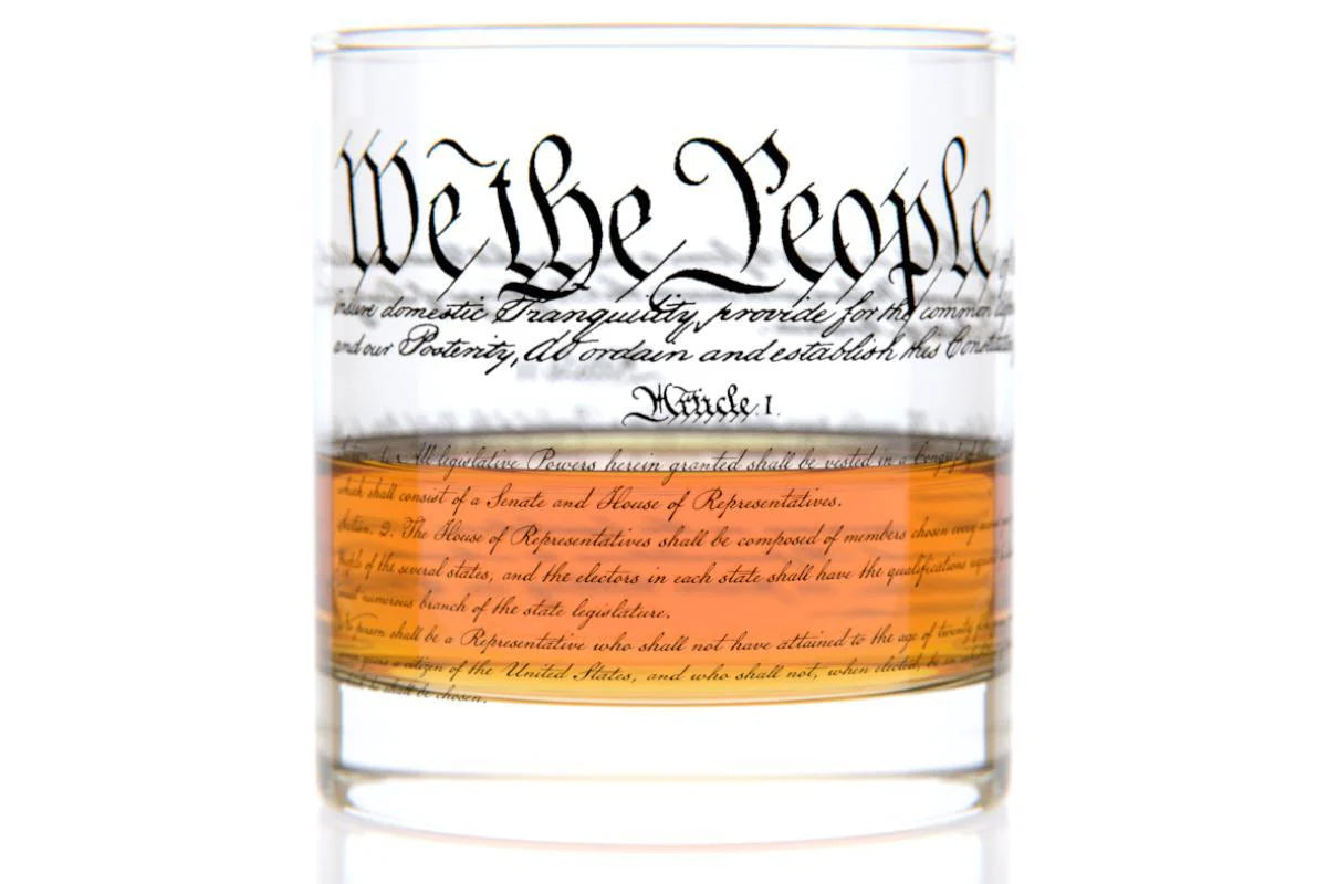 This 11 oz rocks glass is made of clear, heavy glass with a weighted base and a smooth, chip‑resistant rim. Near the top, “We the People” appears in period‑style cursive, while the exterior is wrapped with printed text of the U.S. Constitution. A clear orange band with additional cursive text encircles the lower half of the glass.