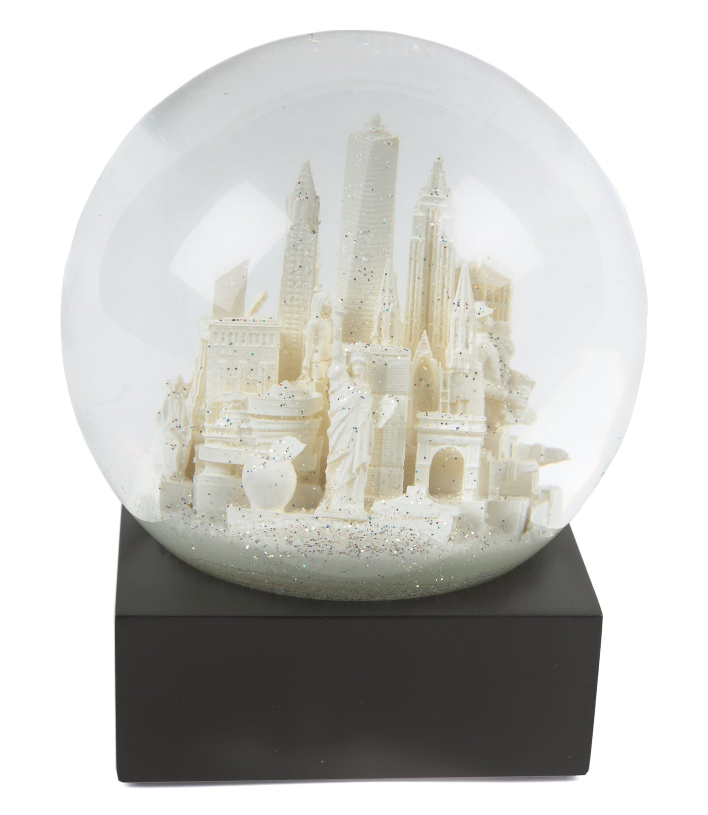 Snow Globe: NYC White —
A round glass snow globe about 4 inches wide and 4.5 inches tall. Inside the clear globe is a small white model of New York City landmarks, including simplified shapes of the Chrysler Building, One World Trade Center, and the Wall Street bull. The buildings are smooth with no raised texture. The globe sits on a solid square black base with flat sides. When shaken, fine white flakes swirl around the miniature skyline. The overall shape is compact.