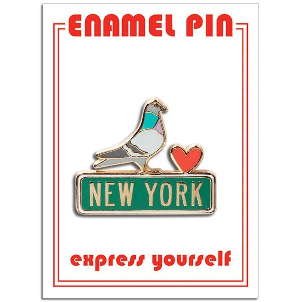 A die‑cut enamel pin showing a stylized pigeon perched on a sign. The pigeon is colored blue, white, gray, and soft mauve, outlined with a thin gold border. Next to it is a small red heart, also edged in gold enamel. The pigeon stands on a green sign that reads “New York” in off‑white block letters.
The pin is attached to rectangular packaging. The backing card is white, with the words “enamel pin” printed in bold red at the top, and the phrase “express yourself” printed near the bottom.