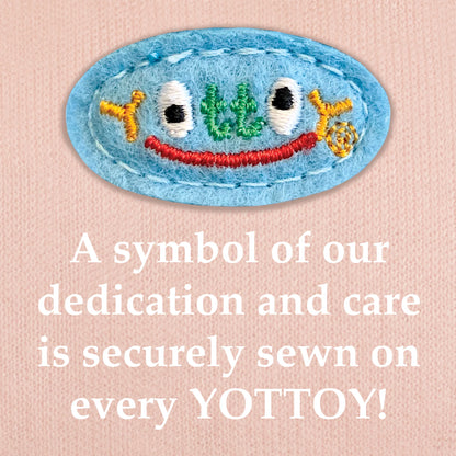 The image shows an embroidered blue YOTTOY logo in multicolored stitching with a small red smile, sewn onto pink knit fabric. Below the emblem, bold white text reads: “A symbol of our dedication and care is securely sewn on every YOTTOY!”