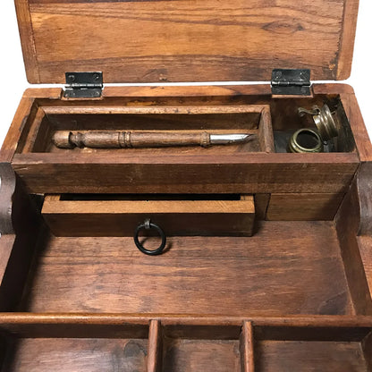 The image shows a weathered, portable teak wood writing desk with its lid open, revealing interior storage compartments. Inside are a vintage‑style wooden pen and a small ink bottle topped with a distressed brass cap, reinforcing the desk’s antique, well‑traveled look.