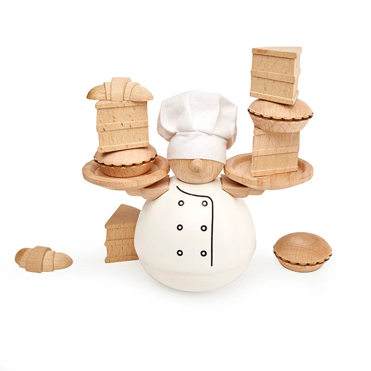Product image of a tabletop stacking game crafted from natural beechwood. At the center is a minimalist wooden baker wearing a white painted outfit and a white fabric chef’s hat, holding small wooden trays in both hands. Balanced precariously across the trays is a growing stack of wooden baked goods, including croissants, cakes, and pies. The warm wood tones, simple design, and unstable tower highlight the game's skill-based balancing challenge against a white background.