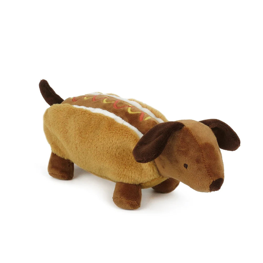 On a plain white background is a Hot Dog Plushie toy shaped like a dachshund. The dog’s body resembles a hot dog: two tan plush buns form the sides, with a darker brown plush hot dog between them. Wavy embroidered lines on top suggest mustard and ketchup. The dog has dark brown floppy ears, short stubby legs, a small tail, and simple stitched details for the eyes and nose. The overall style is soft, playful, and cartoon‑like.