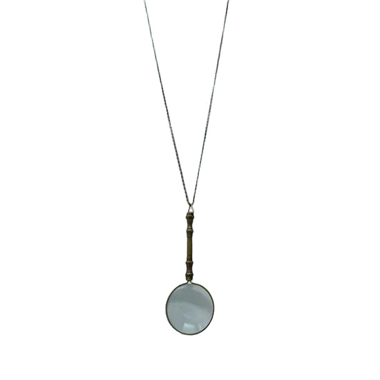 On a white background, the necklace displays an ornate magnifying glass pendant made of aged brass. The magnifying glass is about four inches long, with a decorative handle and a clear, working lens. It hangs from a matching aged brass scroll chain and closes with a small lobster clasp, giving the piece a refined, antique-inspired appearance.