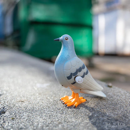 The toy pigeon stands on a real New York curbside, with a green dumpster in the background.