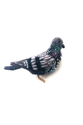 A small pigeon brooch with a simple, elongated body and rounded head. Its surface is richly textured with soft fabric, raised stitching, and beadwork, with sequins adding a gentle shimmer in shades of green, grey, and black. A smooth metal pin is attached to the back for fastening, and each handmade piece features slight variations in texture and detail.