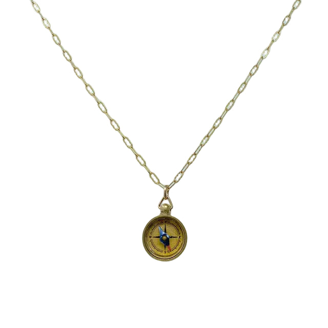 The image shows a delicate pendant necklace with a small, round compass charm about half an inch wide, finished in warm gold‑toned brass. The smooth compass face has lightly engraved directional markings and hangs from a slim, gold‑plated brass cable chain with a small lobster clasp.