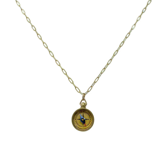 The image shows a delicate pendant necklace with a small, round compass charm about half an inch wide, finished in warm gold‑toned brass. The smooth compass face has lightly engraved directional markings and hangs from a slim, gold‑plated brass cable chain with a small lobster clasp.