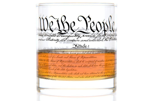 This 11 oz rocks glass is made of clear, heavy glass with a weighted base and a smooth, chip‑resistant rim. Near the top, “We the People” appears in period‑style cursive, while the exterior is wrapped with printed text of the U.S. Constitution. A clear orange band with additional cursive text encircles the lower half of the glass.