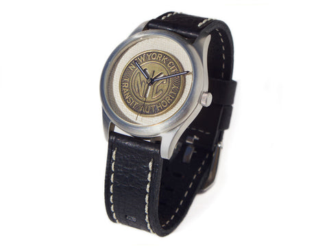 A wristwatch with a thick black leather wrist strap and a bronze NYC subway token as the face.