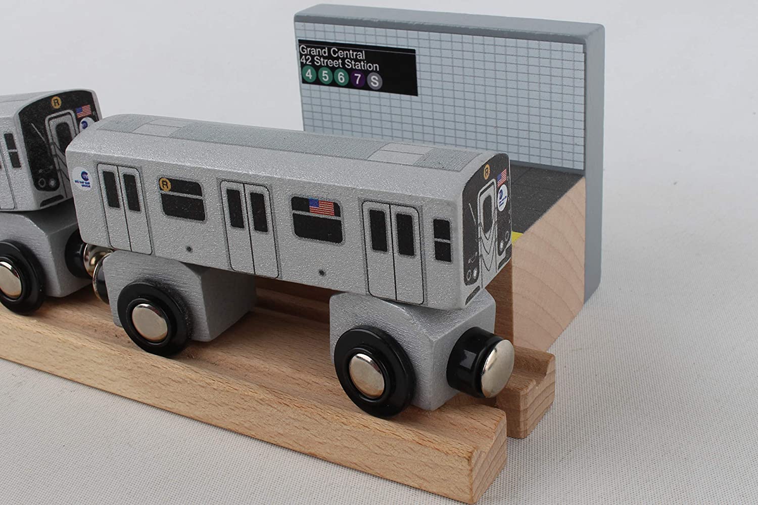 A silver‑gray MTA R‑train subway car sits on wooden tracks beside a black platform with a yellow edge. Behind it is a gray‑blue tiled wall with a sign reading “Grand Central – 42 Street Station,” along with the train line symbols.