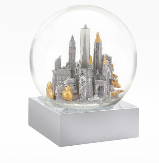 A small, round glass snow globe, about 4 inches wide and 4.5 inches tall, sits firmly on a personalizable silver‑gray square base that feels solid and slightly heavy in the hand. Inside the clear globe is a silver miniature of New York City with subtle gold accents, showing the Chrysler Building, One World Trade Center, and the Wall Street bull. When shaken, fine white flakes softly swirl around the compact skyline.