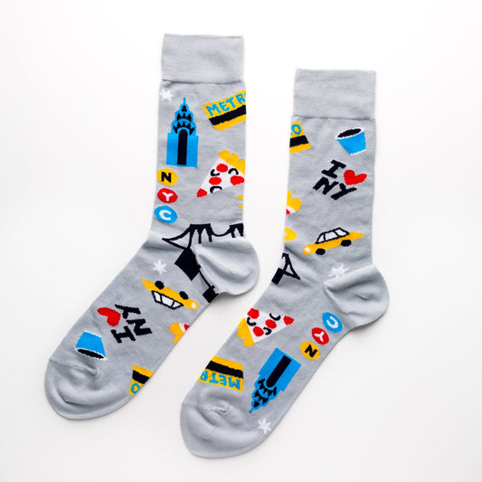 A pair of ultra‑soft cotton crew socks with an all‑over pattern of New York City icons—yellow taxi cabs, pizza slices, a metro card, a coffee cup, the Chrysler Building, and a bridge. The designs are in bright blue, yellow, red, and black on a light grey background, making the imagery stand out clearly.