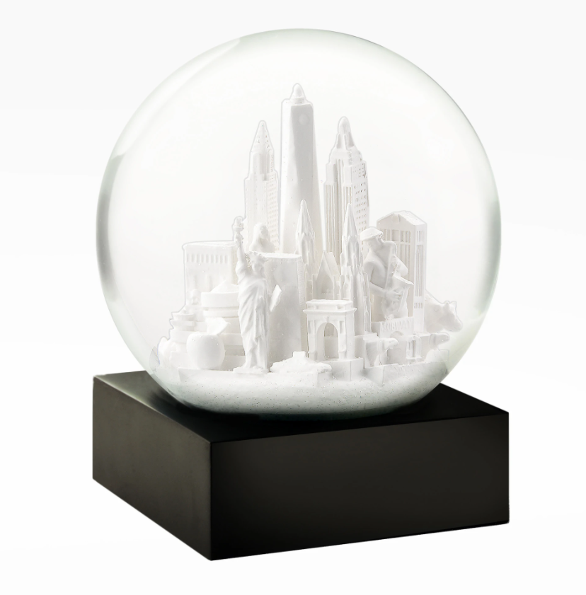 Snow Globe: NYC White — 
A round glass snow globe about 4 inches wide and 4.5 inches tall. Inside the clear globe is a small white model of New York City landmarks, including simplified shapes of the Chrysler Building, One World Trade Center, and the Wall Street bull. The buildings are smooth with no raised texture. The globe sits on a solid square black base with flat sides. When shaken, fine white flakes swirl around the miniature skyline. The overall shape is compact.