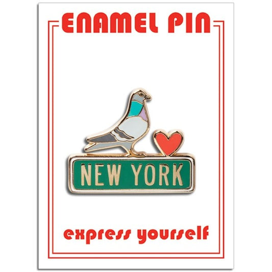 A die‑cut enamel pin showing a stylized pigeon perched on a sign. The pigeon is colored blue, white, gray, and soft mauve, outlined with a thin gold border. Next to it is a small red heart, also edged in gold enamel. The pigeon stands on a green sign that reads “New York” in off‑white block letters.
The pin is attached to rectangular packaging. The backing card is white, with the words “enamel pin” printed in bold red at the top, and the phrase “express yourself” printed near the bottom.